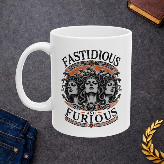 White ceramic mug with a Humorous design combining 'Fast and Furious' with Greek mythology on a white ceramic mug. Text reads 'FASTIDIOUS AND FURIOUS' in distressed lettering surrounding three faces of Medusa in black and white engraving style. Detailed serpents with individual scales form her hair. Greek key pattern borders in orange frame the composition with decorative sun motifs and a small snake head icon at center. Blends classical mythology with contemporary pop culture humor.
