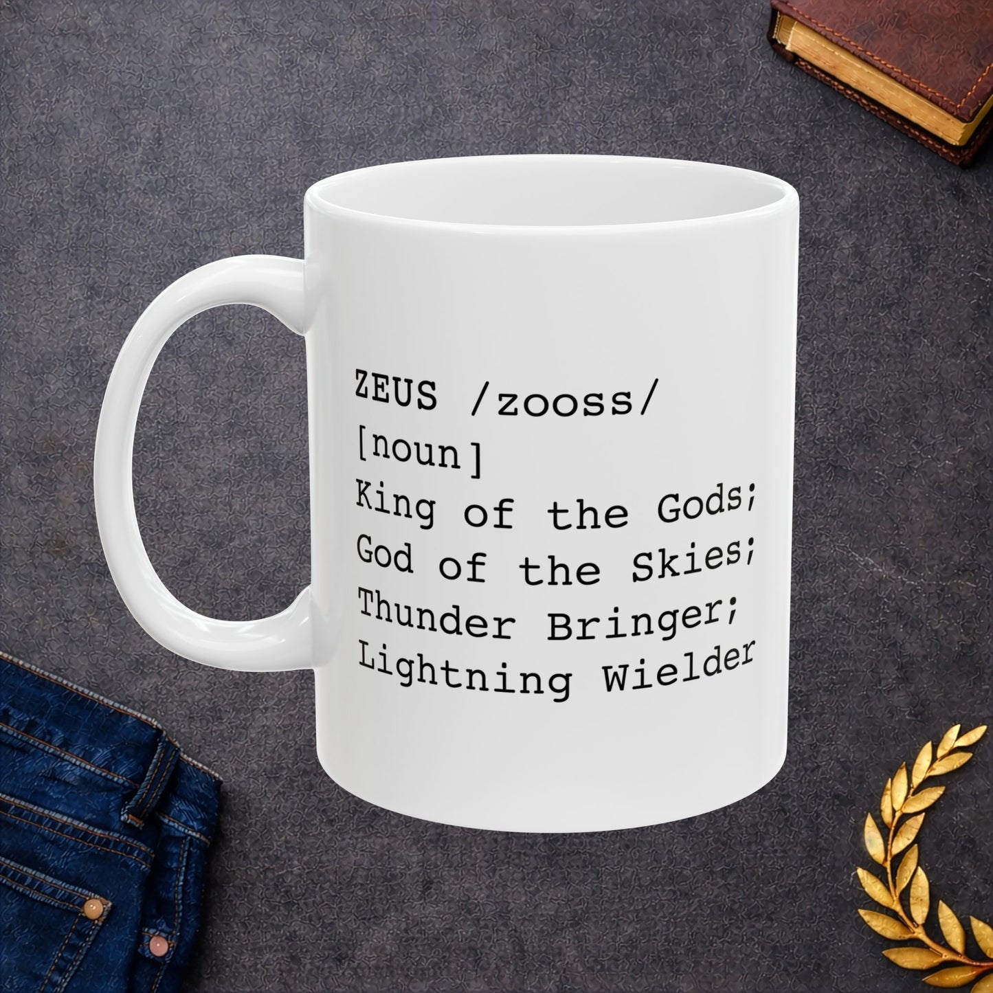 A minimalist black graphic for a white ceramic mug, styled as a dictionary entry. The design features the name "ZEUS" in a bold typewriter font, followed by the phonetic pronunciation "/zooss/," the word class "[noun]," and the definition: "King of the Gods; God of the Skies; Thunder Bringer; Lightning Wielder.