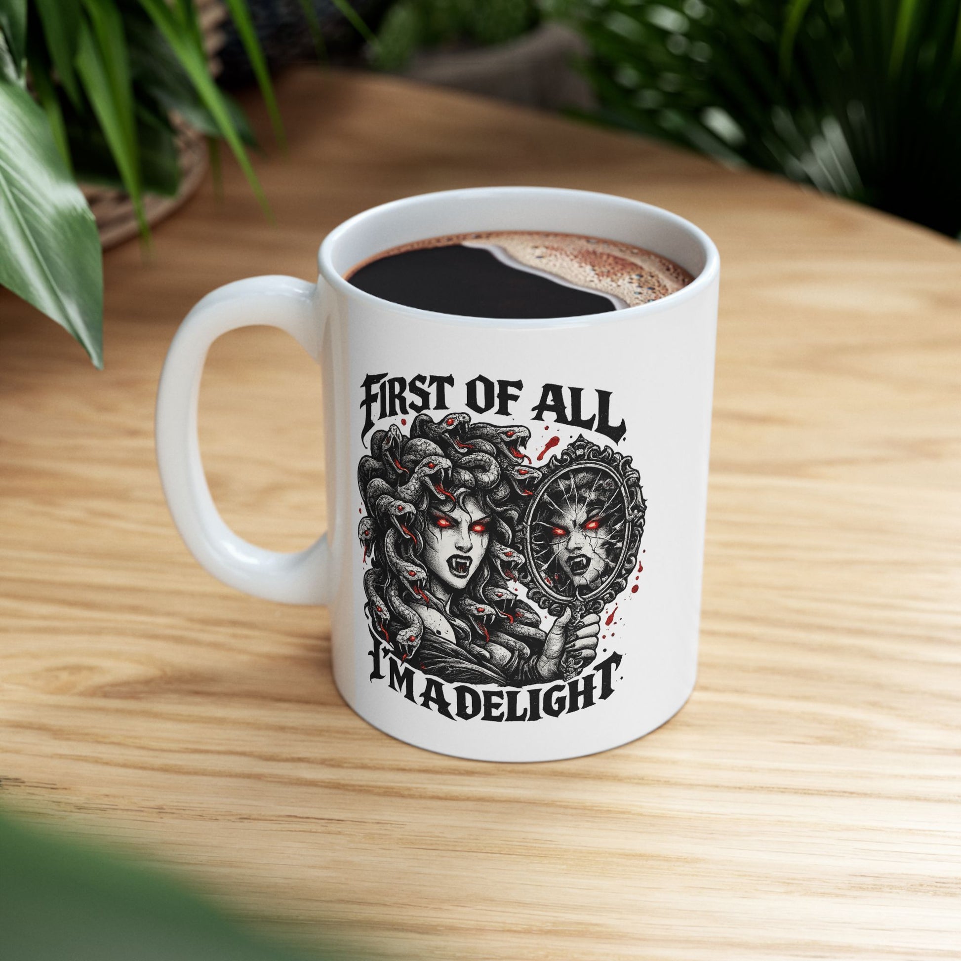 Gothic streetwear illustration of Medusa on a ceramic white mug. Black and white engraving features the figure with serpents for hair, each snake with glowing red eyes. She holds an ornate mirror reflecting her fierce expression with matching red-eyed gaze. Text reads 'First of All, I'm A Delight' in bold metal lettering. Red accents highlight the snakes' eyes. Clever wordplay blends mythology with modern sarcastic humor and confidence.