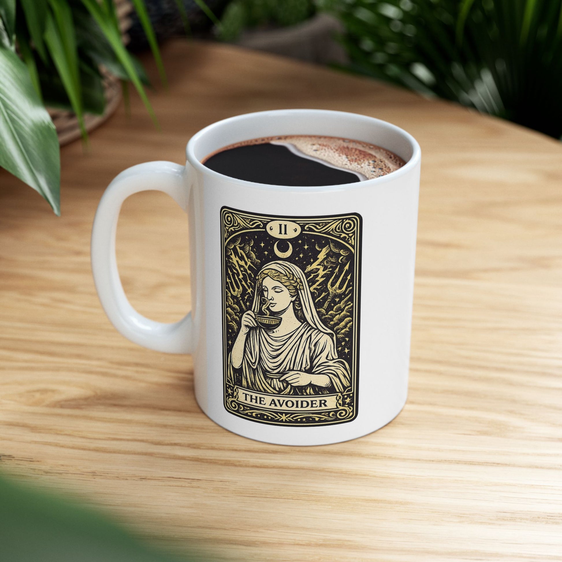 White ceramic mug with a gold and black tarot card-style illustration titled "THE AVOIDER," featuring Hestia from Greek mythology. It depicts the goddess in serene draped robes calmly sipping from a steaming cup, staying unfazed by a chaotic background of stormy clouds, lightning bolts, and tridents. The card includes ornate borders, a crescent moon, and the Roman numeral "II" at the top, creating a humorous, mystical aesthetic.