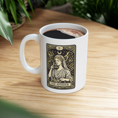 White ceramic mug with a gold and black tarot card-style illustration titled "THE AVOIDER," featuring Hestia from Greek mythology. It depicts the goddess in serene draped robes calmly sipping from a steaming cup, staying unfazed by a chaotic background of stormy clouds, lightning bolts, and tridents. The card includes ornate borders, a crescent moon, and the Roman numeral "II" at the top, creating a humorous, mystical aesthetic.