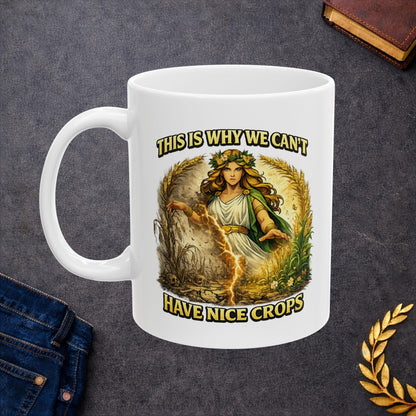 White ceramic mug with a dramatic illustration of Demeter, the Greek goddess of agriculture, standing over a split field. A glowing lightning strike cracks down the center, dividing flourishing, green crops on the right from dead, withered stalks and skulls on the left. Arching text above and below reads "THIS IS WHY WE CAN'T HAVE NICE CROPS" in a bold, weathered yellow font. The design has an intense, mythical comic art style.