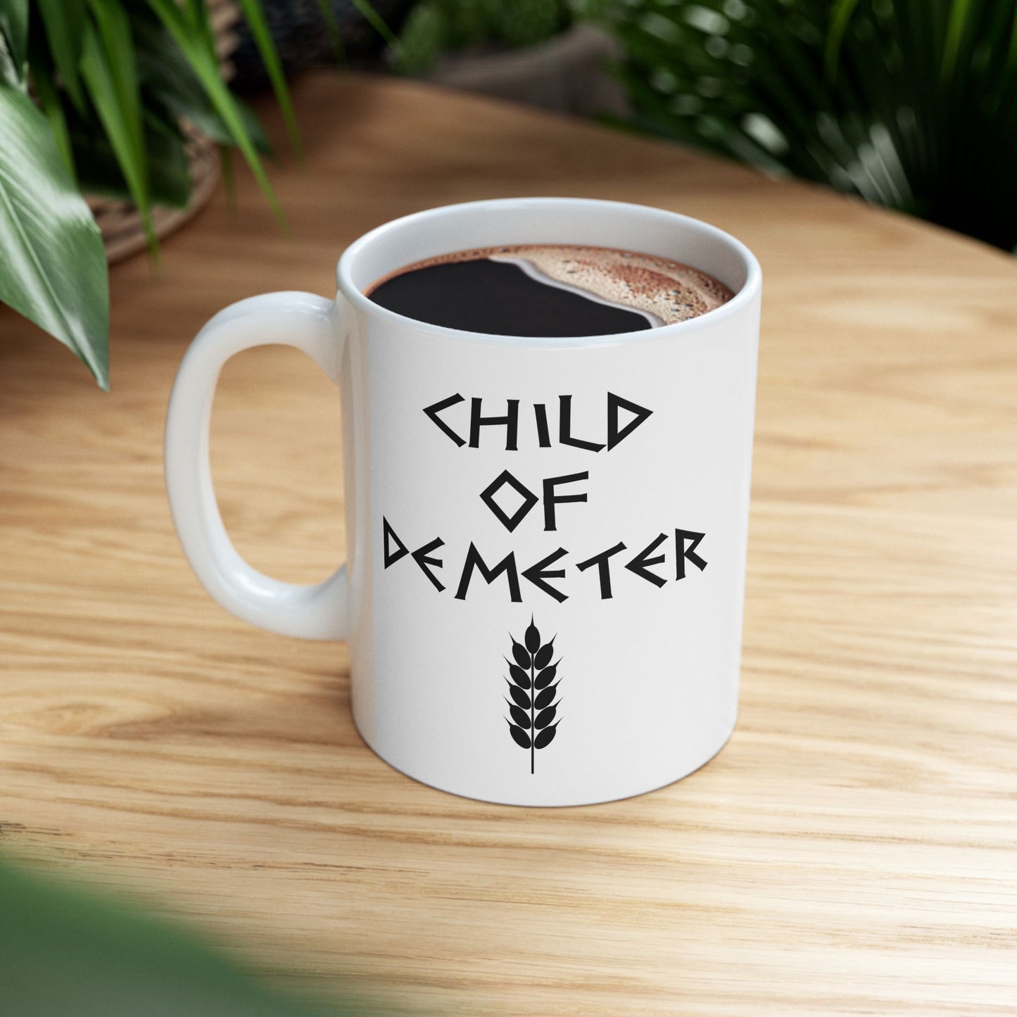 White ceramic mug featuring a minimalist, Greek-inspired graphic in black ink on a white background. The design reads 'CHILD OF DEMETER' in a stylized, angular font, positioned above a clean, line-art graphic of a bundle of harvested wheat.