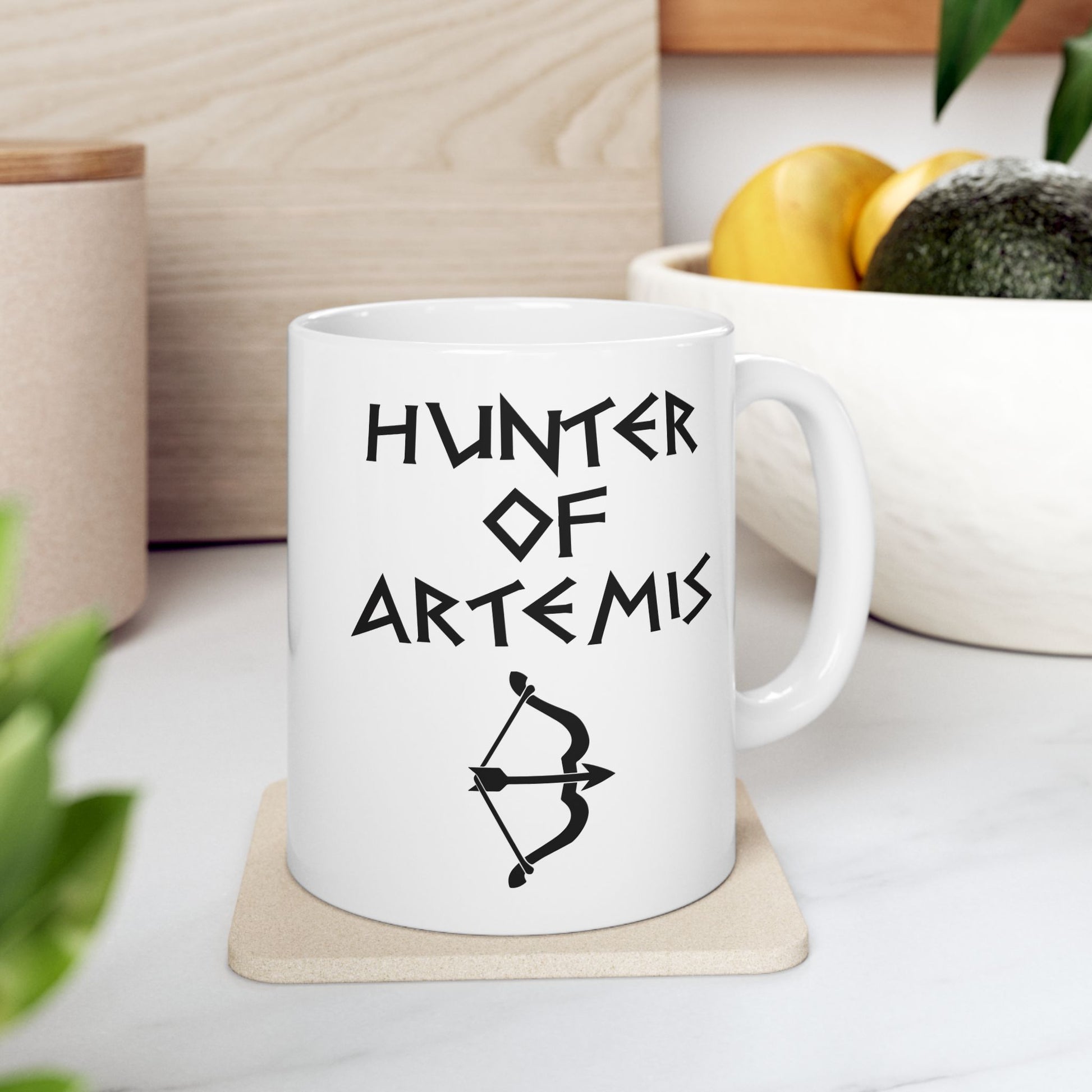 White ceramic mug featuring a minimalist, Greek-inspired graphic in black ink on a white background. The design reads 'HUNTER OF ARTEMIS' in a stylized, angular font, positioned above a clean, line-art graphic of a bow and a notched arrow.