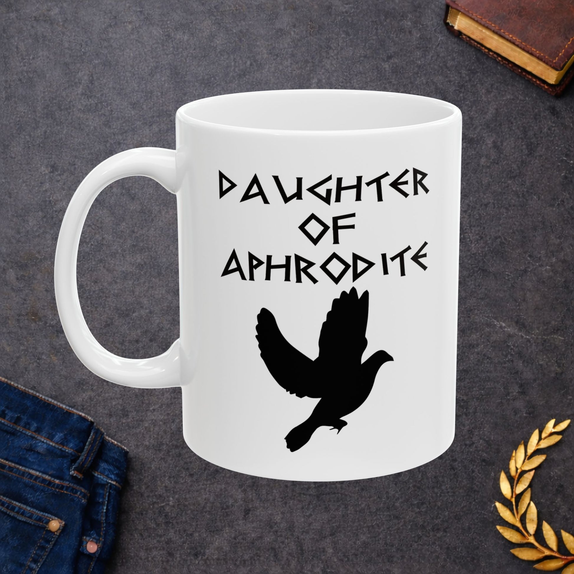White ceramic mug featuring a minimalist, Greek-inspired graphic in black ink on a white background. The design reads 'DAUGHTER OF APHRODITE' in a stylized, angular font, positioned above a clean, line-art graphic of a dove in flight.