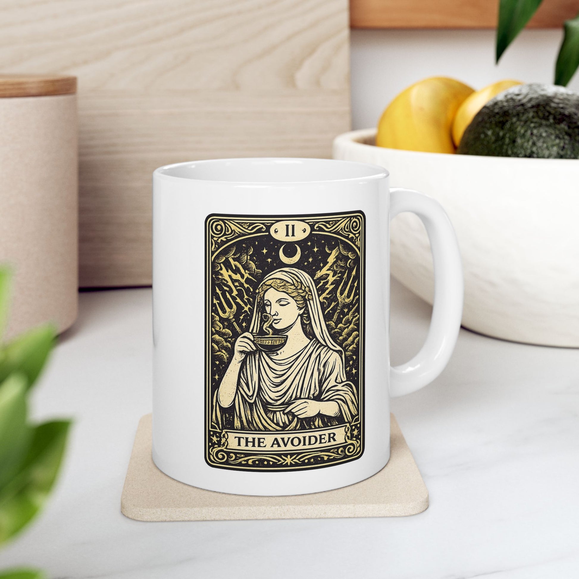 White ceramic mug with a gold and black tarot card-style illustration titled "THE AVOIDER," featuring Hestia from Greek mythology. It depicts the goddess in serene draped robes calmly sipping from a steaming cup, staying unfazed by a chaotic background of stormy clouds, lightning bolts, and tridents. The card includes ornate borders, a crescent moon, and the Roman numeral "II" at the top, creating a humorous, mystical aesthetic.