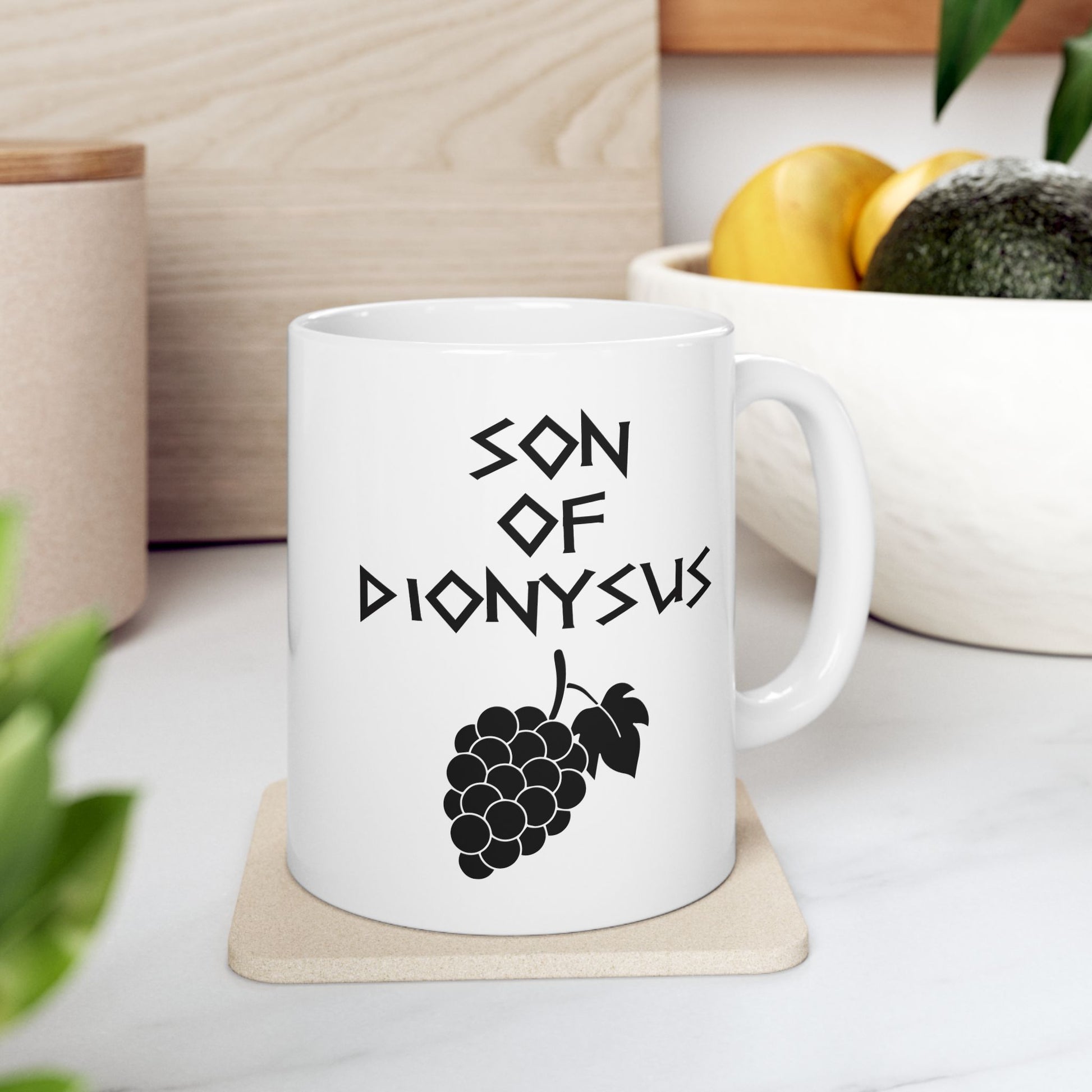 White ceramic mug featuring a minimalist, Greek-inspired graphic in black ink on a white background. The design reads 'SON OF DIONYSUS' in a stylized, angular font, positioned above a clean, line-art graphic of a cluster of grapes hanging from a vine with leaves.