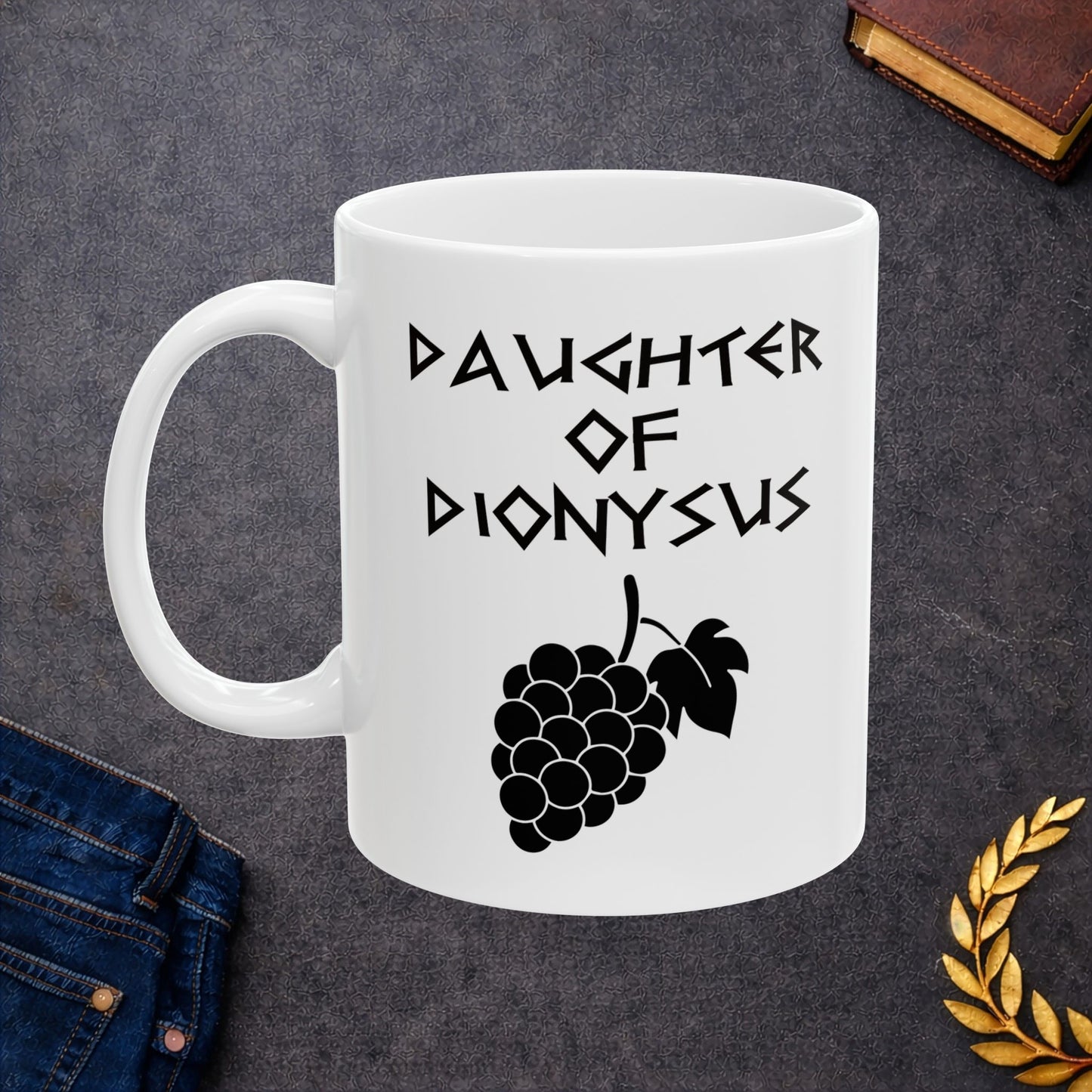 White ceramic mug featuring a minimalist, Greek-inspired graphic in black ink on a white background. The design reads 'DAUGHTER OF DIONYSUS' in a stylized, angular font, positioned above a clean, line-art graphic of a cluster of grapes hanging from a vine with leaves.