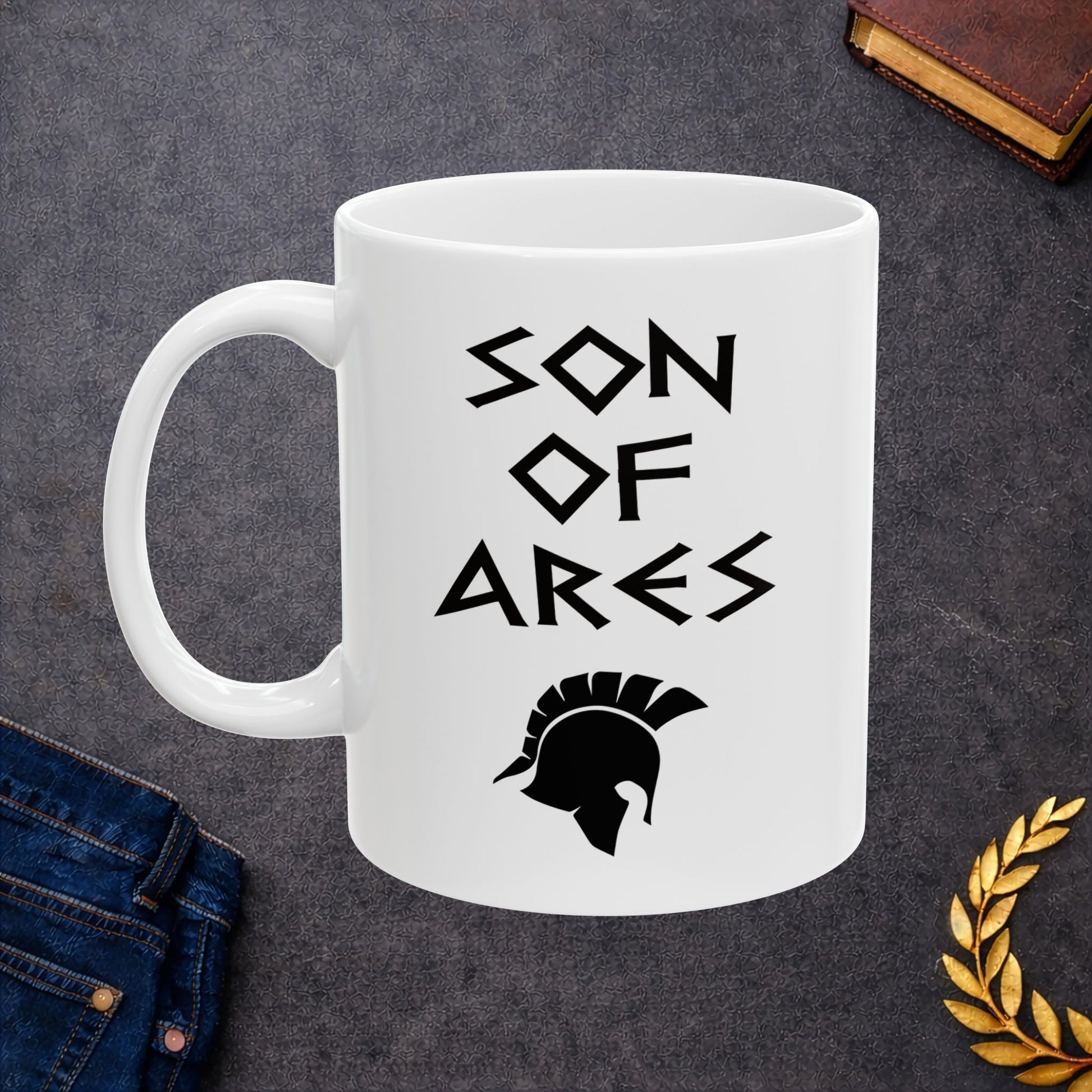White ceramic mug featuring a minimalist, Greek-inspired graphic in black ink on a white background. The design reads 'SON OF ARES' in a stylized, angular font, positioned above a clean, line-art graphic of a Spartan-style plumed warrior helmet.