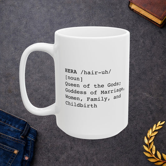 White ceramic mug with a minimalist black text design styled like a dictionary entry. It displays the word "HERA" in capital letters, followed by the pronunciation guide "/hair-uh/" and the part of speech "[noun]". The definition underneath reads: "Queen of the Gods; Goddess of Marriage, Women, Family, and Childbirth." The layout uses a clean, modern typewriter-style font.