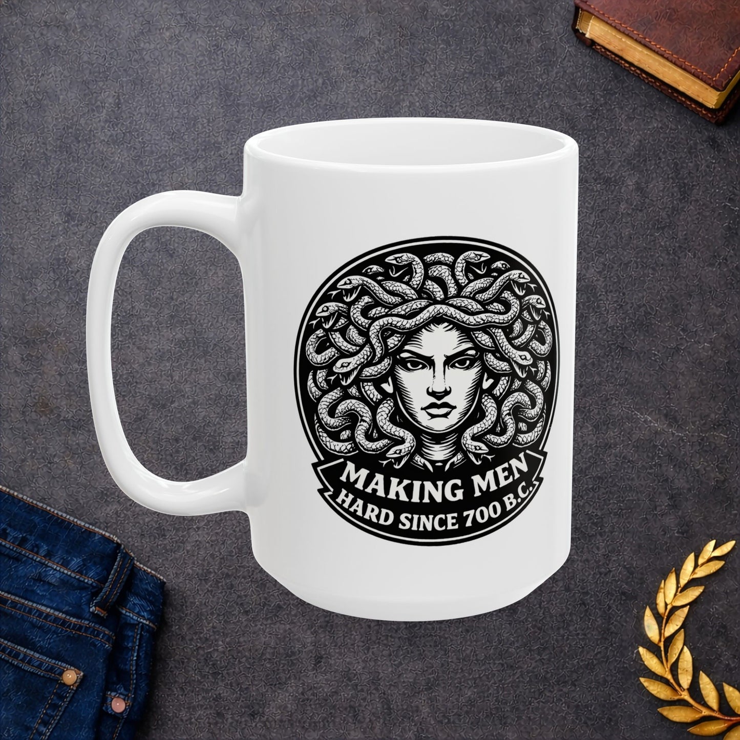 White ceramic mug with a bold black-and-white circular emblem featuring the head of Medusa with a mass of detailed, writhing snakes for hair. Below the illustration, a curved banner contains the humorous double-entendre "MAKING MEN HARD SINCE 700 B.C." written in a heavy, white serif font. The design uses a high-contrast, woodcut-inspired aesthetic with intricate line work against a dark background.