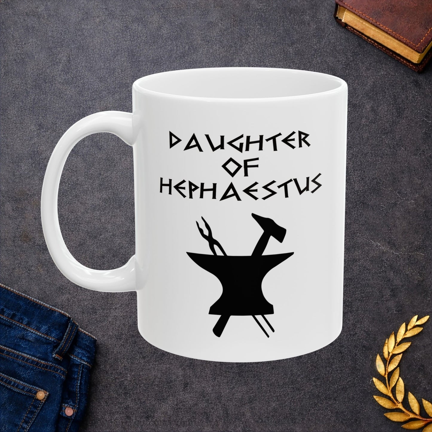 A minimalist black graphic for a white ceramic mug. The design features a central silhouette of a blacksmith's anvil with a hammer and tongs crossed behind it. Above the tools, the text "DAUGHTER OF HEPHAESTUS" is written in a sharp, angular font inspired by ancient Greek inscriptions. The entire illustration is rendered in a clean, solid black style.