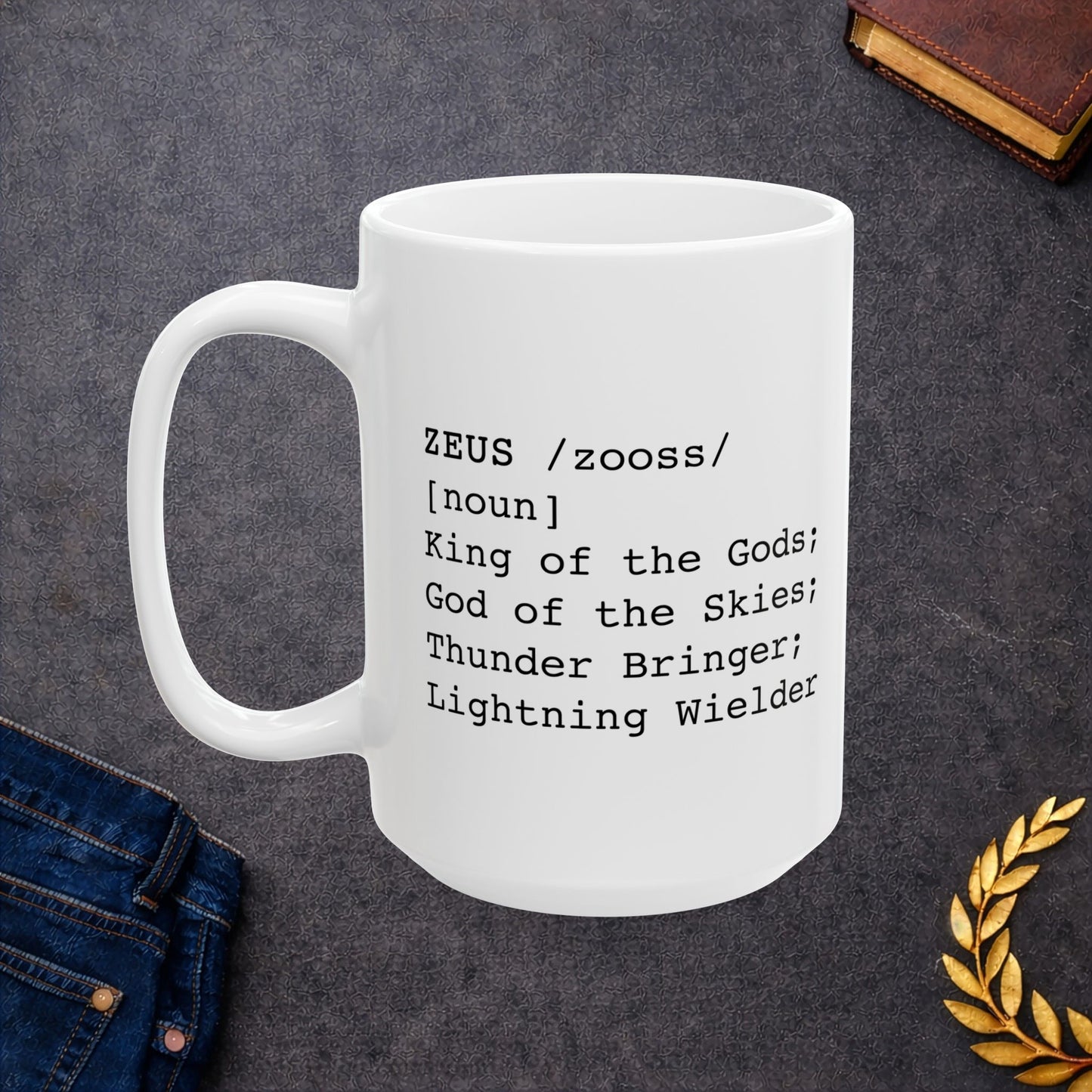 A minimalist black graphic for a white ceramic mug, styled as a dictionary entry. The design features the name "ZEUS" in a bold typewriter font, followed by the phonetic pronunciation "/zooss/," the word class "[noun]," and the definition: "King of the Gods; God of the Skies; Thunder Bringer; Lightning Wielder.