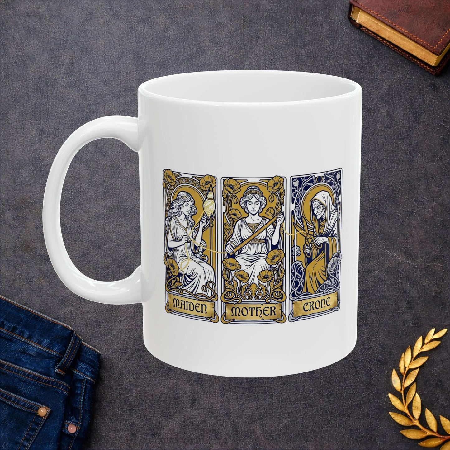 White ceramic mug with Three tarot-style cards depicting the triple goddess archetype: Maiden, Mother, and Crone. The maiden holds a bow surrounded by yellow poppies, the mother wears a crown with a sword, and the crone wears hooded robes with a staff. Each rendered in Art Nouveau style with ornate decorative borders, gold halos, and floral flourishes. Labels appear at card bases. Celebrates the feminine divine in all life stages and cyclical transformation.