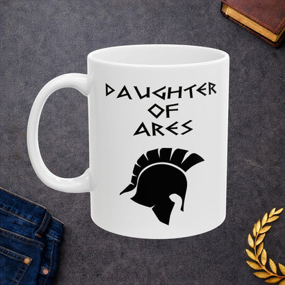 White ceramic mug featuring a minimalist, Greek-inspired graphic in black ink on a white background. The design reads 'DAUGHTER OF ARES' in a stylized, angular font, positioned above a clean, line-art graphic of a Spartan-style plumed warrior helmet.