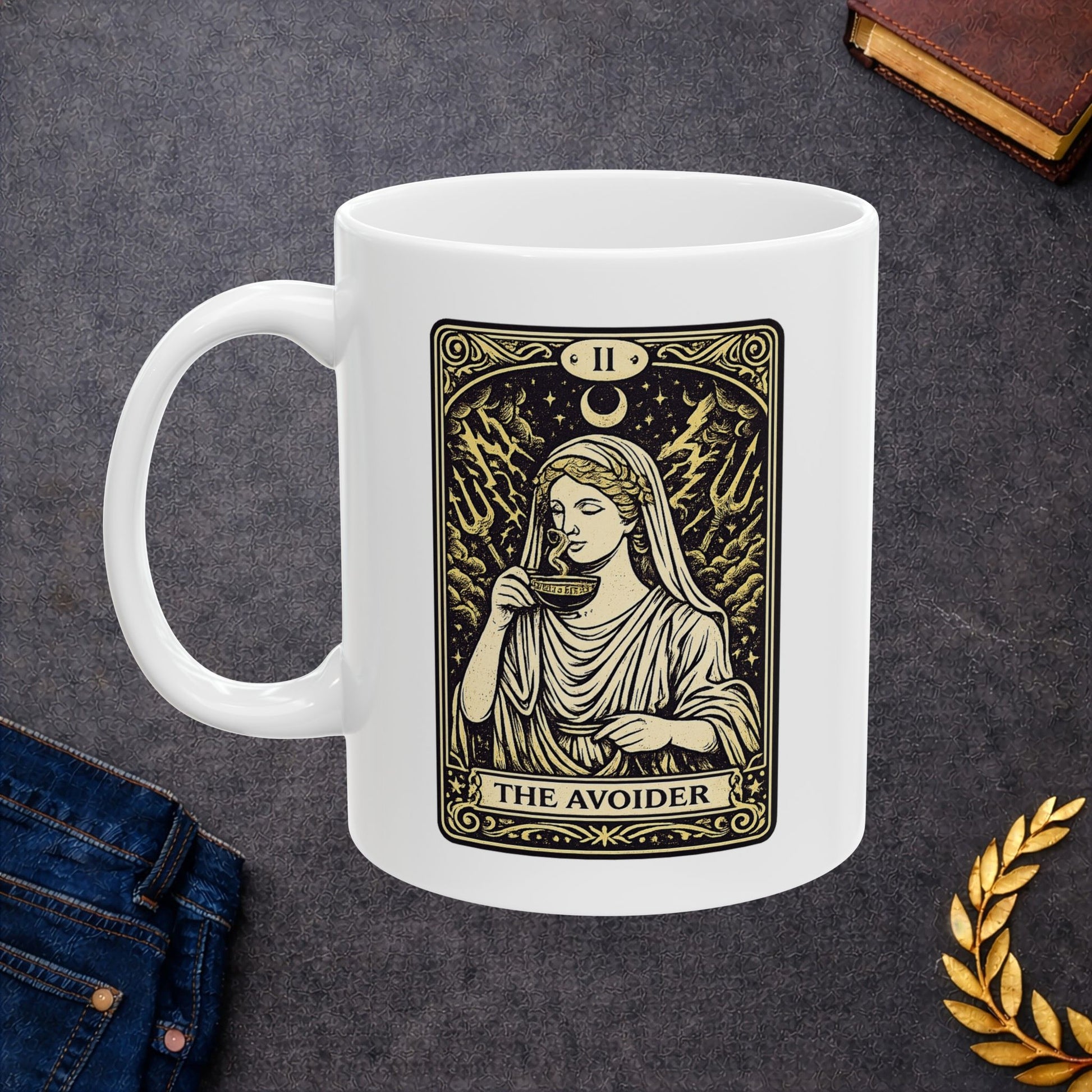 White ceramic mug with a gold and black tarot card-style illustration titled "THE AVOIDER," featuring Hestia from Greek mythology. It depicts the goddess in serene draped robes calmly sipping from a steaming cup, staying unfazed by a chaotic background of stormy clouds, lightning bolts, and tridents. The card includes ornate borders, a crescent moon, and the Roman numeral "II" at the top, creating a humorous, mystical aesthetic.