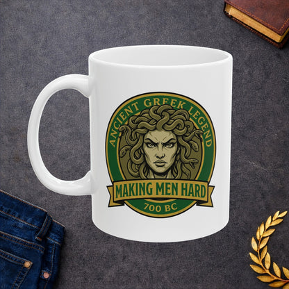 Funny Greek mythology mug design featuring a bold vintage badge with Medusa's stern face surrounded by writhing snakes. The circular emblem reads "Ancient Greek Legend" across the top and "Making Men Hard" on a banner below, with "700 BC" at the bottom. The design uses classic green, gold, and olive tones in a retro emblem style, combining mythology with humorous wordplay.