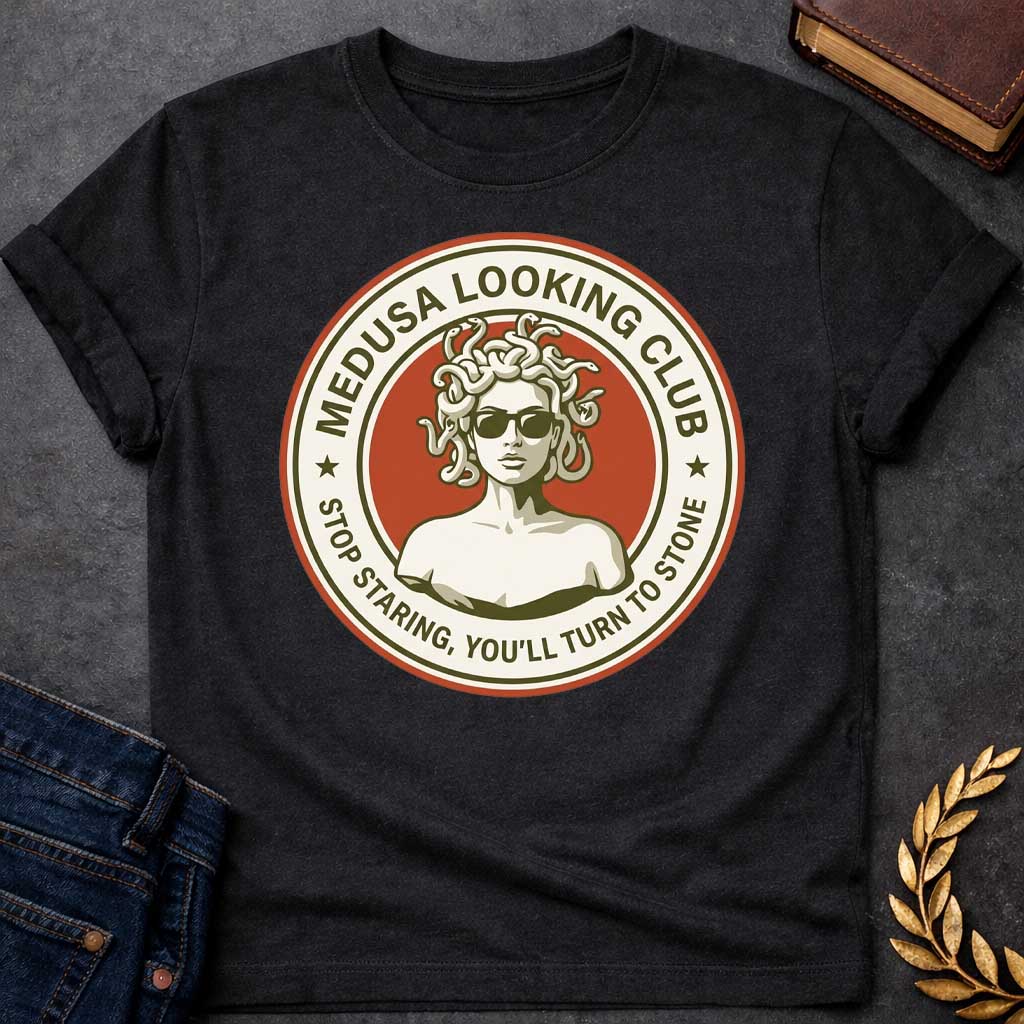 Black t-shirt in retro badge style: a cool, sunglasses-wearing Medusa with snake hair stands confidently against a burnt-orange circle. Cream and olive-green ring text reads "Medusa Looking Club" above and "Stop Staring, You'll Turn to Stone" below, with two stars flanking the design.