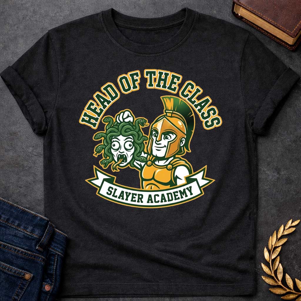 Black t-shirt in collegiate mascot style: a smirking gold-helmeted Greek warrior holds up Medusa's shocked, snarling severed head by her green snake hair. Bold green and gold text arcs above the design, with "Slayer Academy" on a white ribbon banner below. Playful varsity-logo aesthetic on a white background.