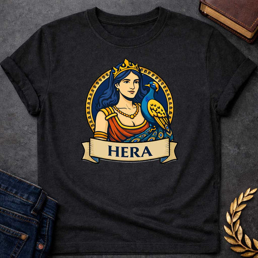 Black T-shirt graphic featuring a vibrant illustrated portrait of Hera, the Greek Queen of the Gods, with long dark blue wavy hair and a gold crown, wearing a red draped robe with gold arm bands and a gold beaded necklace, with a blue and gold peacock perched at her side displaying its ornate tail feathers. She is framed within a gold circular border against a navy background, with a scroll banner below reading "Hera," in a bold tattoo-inspired style with navy, gold, and red tones