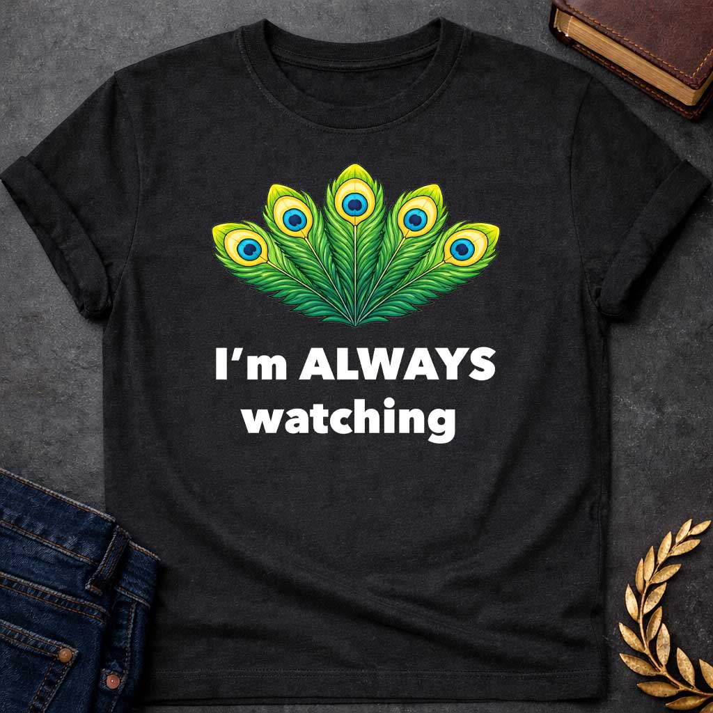 Black T-shirt graphic featuring a vibrant illustrated fan of five peacock feathers in rich green, yellow, and blue tones, each with a distinctive eye-spot, above text reading "I'm Always Watching" — a humorous reference to Hera, the Greek Queen of the Gods, whose sacred symbol is the peacock