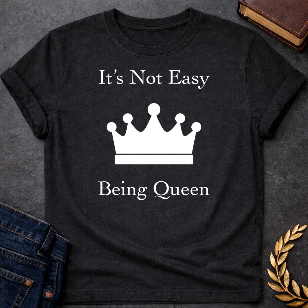 Black T-shirt graphic in white on black featuring elegant serif text reading "It's Not Easy Being Queen" split above and below a simple white crown silhouette icon, in a humorous nod to Hera, the Greek Queen of the Gods