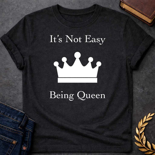 Black T-shirt graphic in white on black featuring elegant serif text reading "It's Not Easy Being Queen" split above and below a simple white crown silhouette icon, in a humorous nod to Hera, the Greek Queen of the Gods