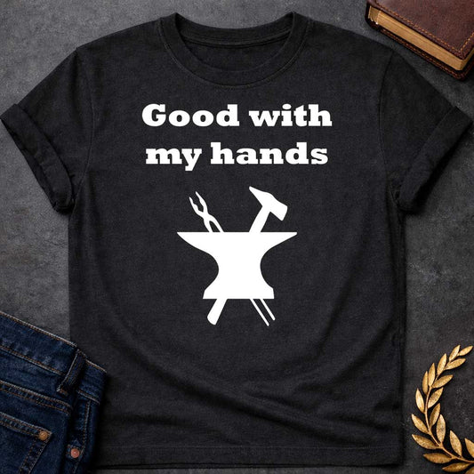 Black T-shirt graphic in white featuring bold serif text reading "Good with my hands" above a flat black silhouette illustration of a blacksmith's anvil with a hammer and tongs crossed on top — a humorous nod to Hephaestus, the Greek God of the Forge