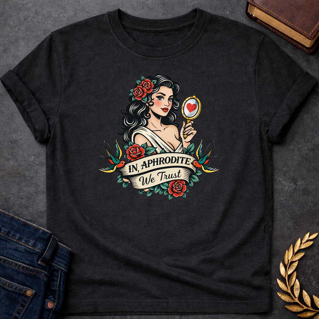 Black T-shirt graphic in a classic American traditional tattoo style featuring a pin-up illustration of Aphrodite, the Greek Goddess of Love, with dark wavy hair adorned with red roses, wearing a white draped gown, holding a gold hand mirror reflecting a red heart. She is flanked by two swooping swallows and surrounded by red roses and green leaves, with a scroll banner below reading "In Aphrodite We Trust" in bold and script lettering