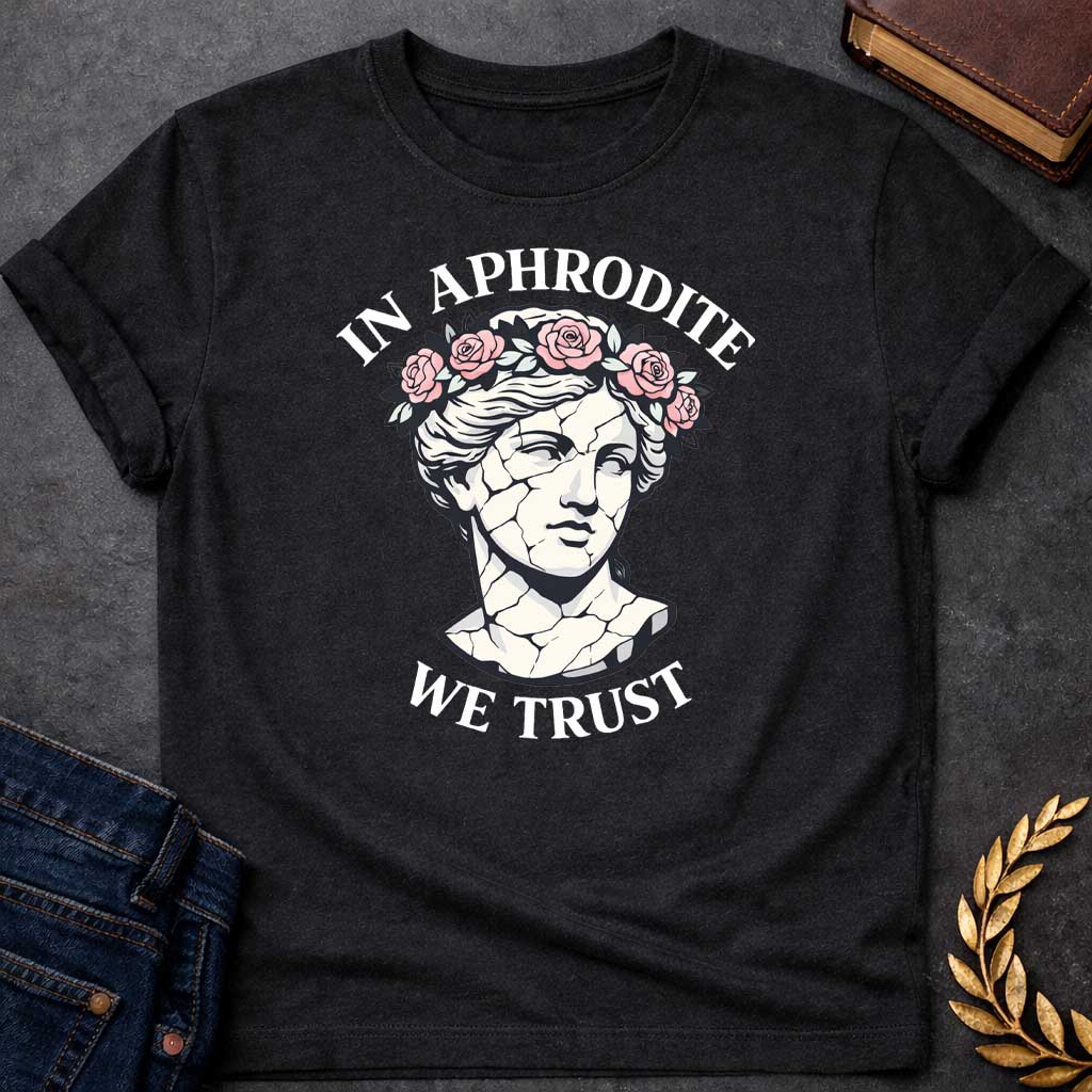 Black T-shirt graphic featuring a cracked marble bust illustration of Aphrodite, the Greek Goddess of Love, adorned with a floral crown of pink roses and green leaves, with bold arched text reading "In Aphrodite" curved above and "We Trust" curved below, in a modern aesthetic vaporwave-inspired style with white text and soft pink and white tones
