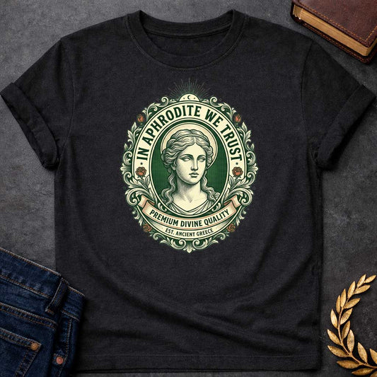 Black T-shirt graphic featuring a vintage apothecary label-style oval badge design in forest green, cream, and terracotta. A detailed engraving-style portrait of Aphrodite is centered within the badge, surrounded by ornate scrollwork and flanked by red roses, with a crescent moon and sunburst radiating from the top. Text arched around the inner circle reads "In Aphrodite We Trust," with a scroll banner below reading "Premium Divine Quality" and smaller text underneath reading "Est. Ancient Greece,"