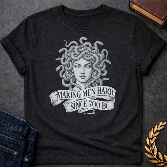 Black T-shirt graphic in grey and white featuring a highly detailed fine-line tattoo-style front-facing portrait of Medusa with numerous coiling snakes for hair, rendered in intricate stippling and engraving technique, with a flowing ribbon banner below reading "Making Men Hard Since 700 BC" — a witty double entendre referencing Medusa's mythological power to turn men to stone