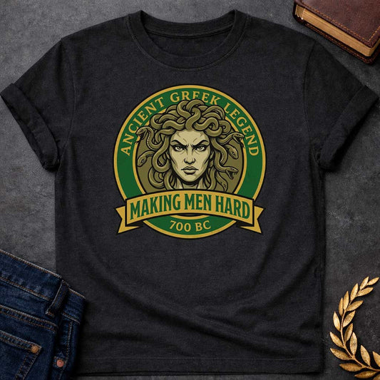 Black T-shirt graphic featuring a bold vintage badge design in forest green and gold. A fierce front-facing illustration of Medusa with coiling olive-toned snakes for hair and an intense glaring expression fills the circular center, with gold arched text reading "Ancient Greek Legend" around the upper border. A gold ribbon banner below the portrait reads "Making Men Hard" in green text, with "700 BC" below it — a witty double entendre referencing Medusa's mythological power to turn men to stone