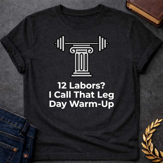 Black T-shirt with a graphic design. At the top, there is a gray illustration of a classical Greek column supporting a barbell with weight plates on both sides. Below the illustration, the white text reads: "12 Labors? I Call That Leg Day Warm-Up"