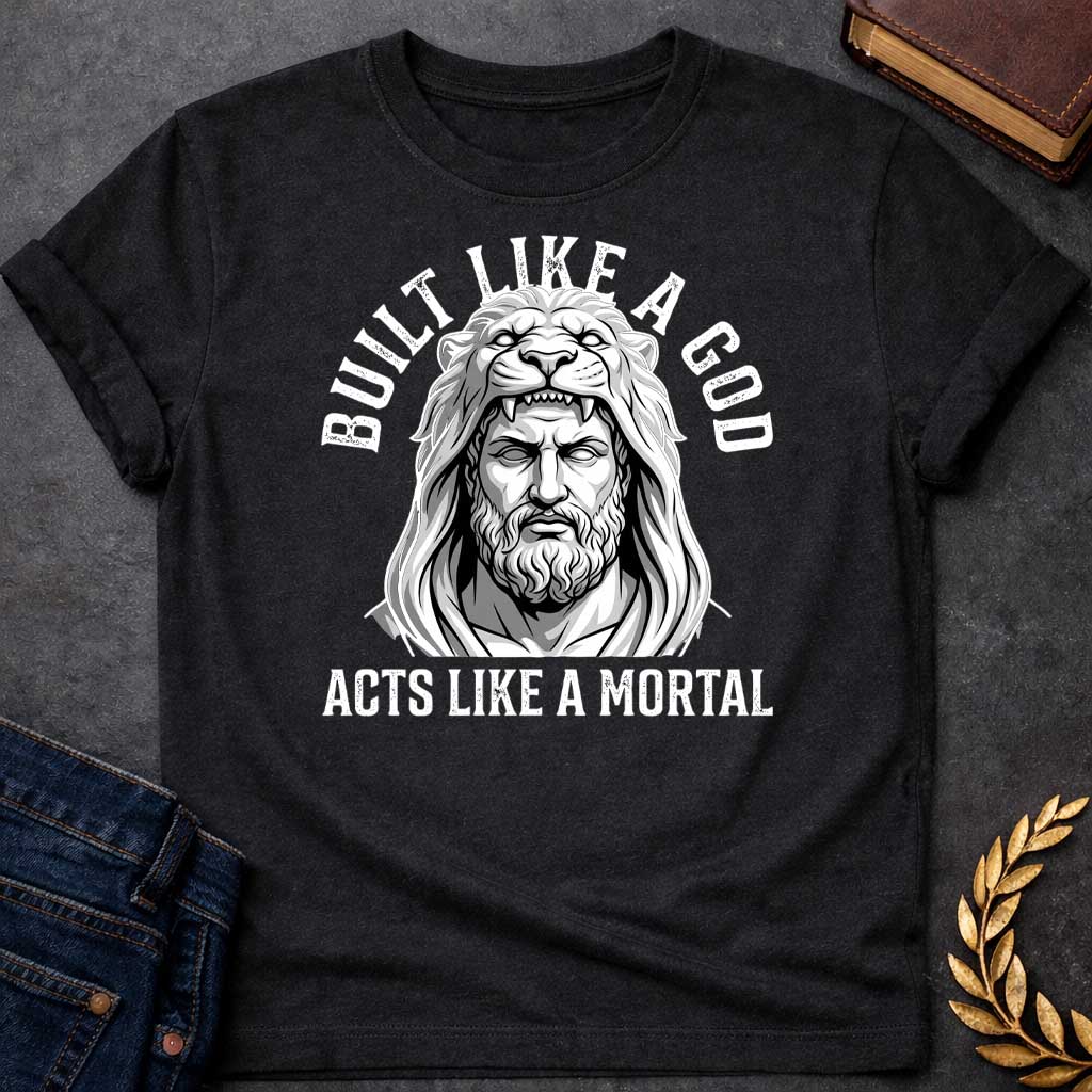 Black T-Shirt with a high-contrast, black-and-white graphic. The illustration depicts a stylized, statuesque man wearing the head and mane of a lion as a hood, reminiscent of Hercules. The text "BUILT LIKE A GOD" is arched over the top, and "ACTS LIKE A MORTAL" is printed in bold, straight text across the botto