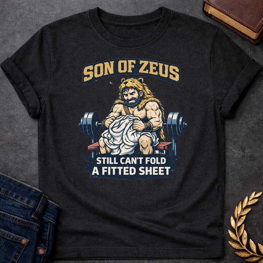 Black T-shirt featuring a humorous illustration of a muscular, bearded man wearing a lion pelt—resembling Hercules—sitting on a weight bench. He is struggling, with a frustrated expression, to fold a crumpled white fitted sheet. Behind him is a barbell. The text at the top reads "SON OF ZEUS" in a distressed, arched font, and the text at the bottom reads "STILL CAN'T FOLD A FITTED SHEET" in a banner