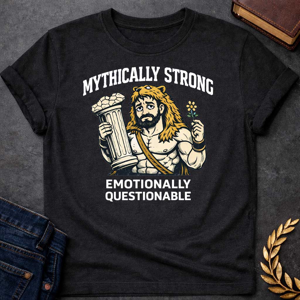 Black T-shirt featuring a humorous illustration of a muscular, bearded man wearing a lion-skin hood—reminiscent of Hercules—with a sad, weary expression. He holds a broken classical column in one hand and a small flower in the other. Arched over the top is the white text "MYTHICALLY STRONG," and below the illustration is the text "EMOTIONALLY QUESTIONABLE,"