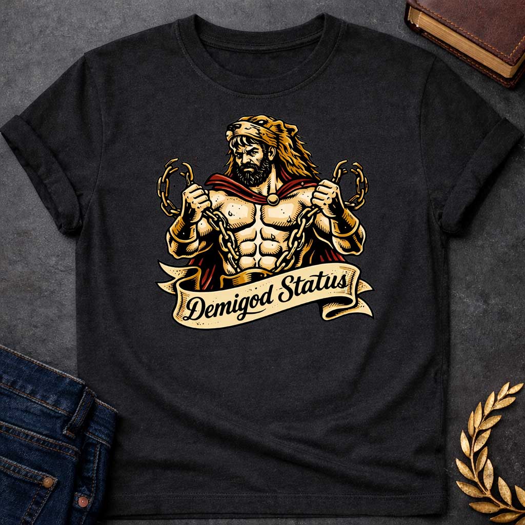 Black T-shirt featuring a muscular, bearded figure resembling Heracles, wearing a lion-skin hood and a red cape, as he heroically snaps thick golden chains with his hands. Below the illustration is a flowing, decorative banner with the text "Demigod Status" written in a stylish script font,