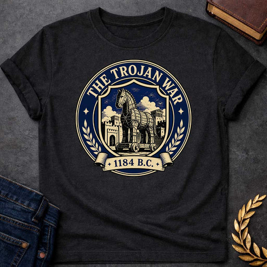 Black T-shirt with a graphic featuring a formal vintage seal design in navy and cream. A detailed engraving-style illustration of the Trojan Horse on wheels is centered within a shield, set against the walled city of Troy with Greek temple columns and dramatic clouds. Text arched around the outer circle reads "The Trojan War" with gold diamond stars, flanked by cream laurel branches, and a scroll banner below reads "1184 B.C." — referencing the legendary Trojan War from Greek mythology