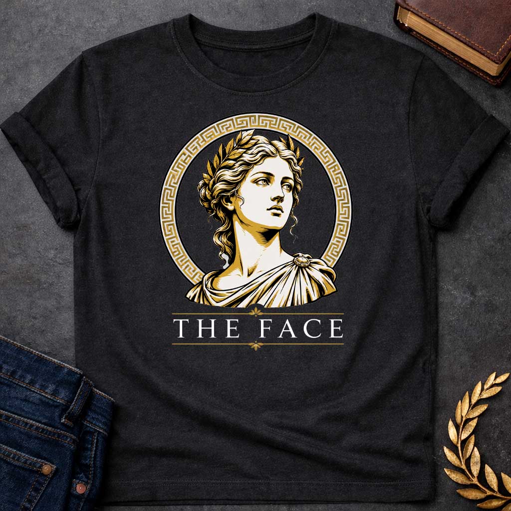 Black T-shirt with a classical Greek graphic featuring a gold and cream vector portrait of Helen of Troy in profile. She wears a golden laurel wreath and a traditional draped chiton, looking upward with a serene expression. The portrait is framed by a circular border with a gold Greek key (meander) pattern. Below the emblem, the text "THE FACE" is written in a clean, elegant white serif font between horizontal gold accent lines.