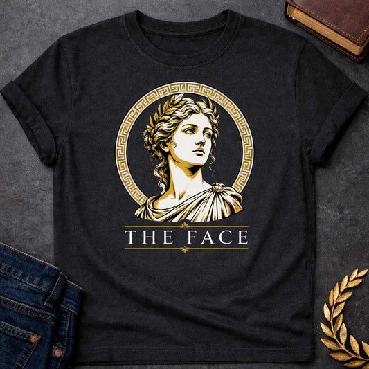 Black T-shirt with a classical Greek graphic featuring a gold and cream vector portrait of Helen of Troy in profile. She wears a golden laurel wreath and a traditional draped chiton, looking upward with a serene expression. The portrait is framed by a circular border with a gold Greek key (meander) pattern. Below the emblem, the text "THE FACE" is written in a clean, elegant white serif font between horizontal gold accent lines.