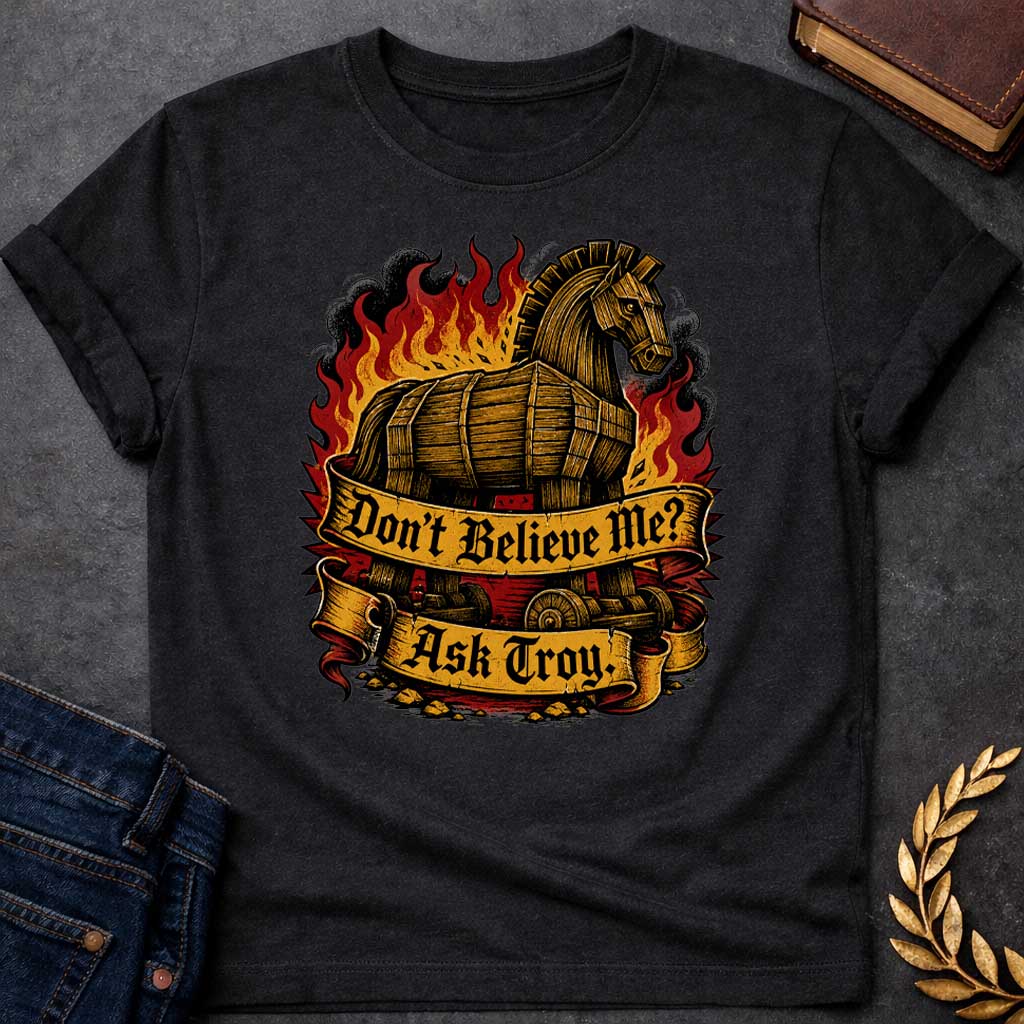 Black T-shirt with a bold illustration of the Trojan Horse engulfed in flames with red, orange, and yellow fire and black smoke. The wooden horse emerges from flames with detailed wood-grain texture. Two ornate gold banners with blackletter text read 'Don't Believe Me?' and 'Ask Troy.' Dark red background with rubble scattered below. References Cassandra's ignored warning about the Trojan Horse that led to Troy's destruction. Dramatic tattoo-inspired style with vintage engraving detail.