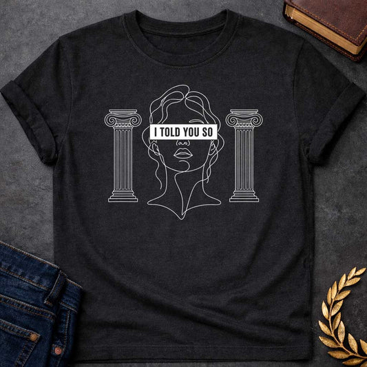 Black T-shirt with a minimalist line art design featuring a continuous white ink sketch of Cassandra or a Greek goddess. Her eyes are covered by a bold, solid black rectangular bar with the text "I TOLD YOU SO" in white sans-serif capital letters. The portrait is flanked on both sides by simple, symmetrical line-drawn Ionic Greek columns.
