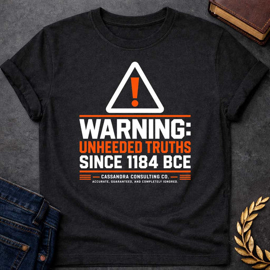 Black T-shirt with a corporate-style satirical design mimicking an industrial hazard sign. At the top is a triangular warning icon with an orange exclamation point. Bold block text reads "WARNING: UNHEEDED TRUTHS SINCE 1184 BCE" in alternating white and orange. Below, it features a faux company name: "CASSANDRA CONSULTING CO." followed by the tagline, "ACCURATE, GUARANTEED, AND COMPLETELY IGNORED."