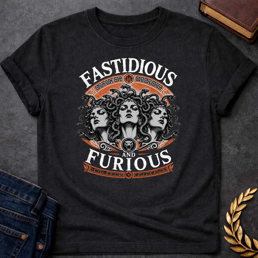 Black T-shirt with a Humorous design combining 'Fast and Furious' with Greek mythology. Text reads 'FASTIDIOUS AND FURIOUS' in distressed lettering surrounding three faces of Medusa in black and white engraving style. Detailed serpents with individual scales form her hair. Greek key pattern borders in orange frame the composition with decorative sun motifs and a small snake head icon at center. Blends classical mythology with contemporary pop culture humor and clever wordplay.