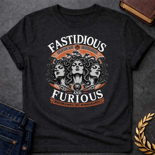 Black T-shirt with a Humorous design combining 'Fast and Furious' with Greek mythology. Text reads 'FASTIDIOUS AND FURIOUS' in distressed lettering surrounding three faces of Medusa in black and white engraving style. Detailed serpents with individual scales form her hair. Greek key pattern borders in orange frame the composition with decorative sun motifs and a small snake head icon at center. Blends classical mythology with contemporary pop culture humor and clever wordplay.