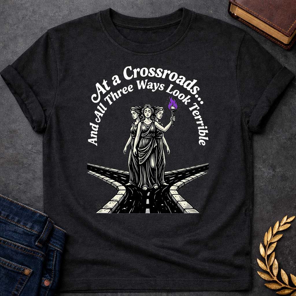 Black T-shirt with an Illustration of the Hecate at a crossroads, rendered in black and white engraving style. The figures wear flowing robes and stand together, with the center holding a flaming torch with purple flames. Text reads 'At a Crossroads: And All Three Ways Look Terrible,' humorously referencing difficult choices and Hecate’s role as goddess of the crossroads. Multiple roads branch beneath them in perspective, receding into darkness symbolizing uncertainty.