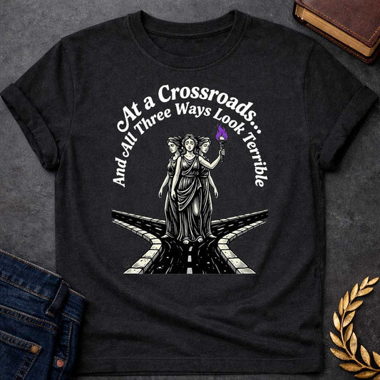 Black T-shirt with an Illustration of the Hecate at a crossroads, rendered in black and white engraving style. The figures wear flowing robes and stand together, with the center holding a flaming torch with purple flames. Text reads 'At a Crossroads: And All Three Ways Look Terrible,' humorously referencing difficult choices and Hecate’s role as goddess of the crossroads. Multiple roads branch beneath them in perspective, receding into darkness symbolizing uncertainty.