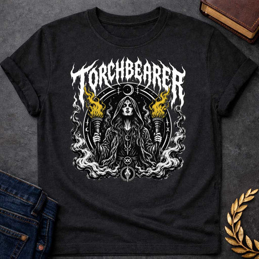 Black T-shirt with a Dark metal band aesthetic illustration titled 'Torchbearer.' Hecate holds two ornate torches with gold and black flames. She wears mystical symbols including a crescent moon necklace and a circular mandala halo with occult iconography. Swirling smoke and flame designs surround the figure in black, white, and gold. Blends heavy metal imagery with Greek mythology and mystical symbolism celebrating enlightenment, divine knowledge, and dark power.