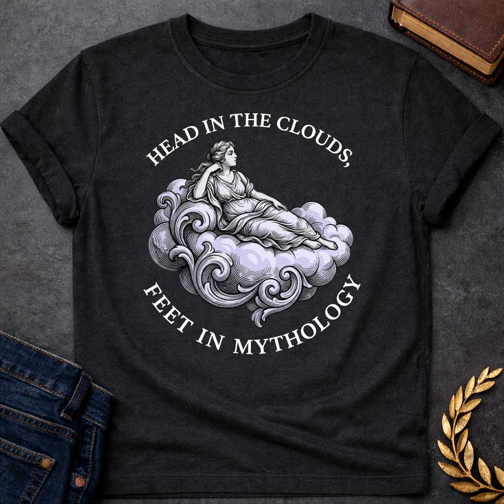 Black T-shirt with a classical Greek mythology t-shirt design featuring a detailed engraving-style illustration. A woman in a traditional draped chiton relaxes gracefully, reclining on a large, swirling baroque-style cloud shaded in a pale purple hue. Arched above her is the text "HEAD IN THE CLOUDS," and curved inversely below the cloud is "FEET IN MYTHOLOGY," all written in a clean white serif font.